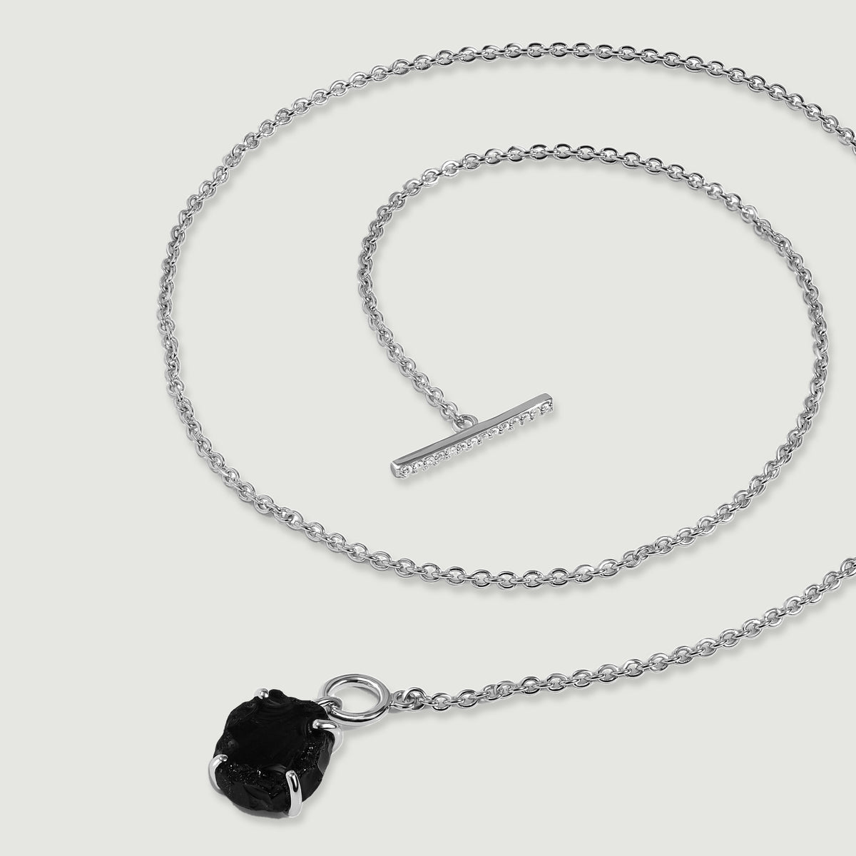 A silver necklace lies in loose curves with its crystal-set bar separated from the circular clasp beside a black obsidian pendant.