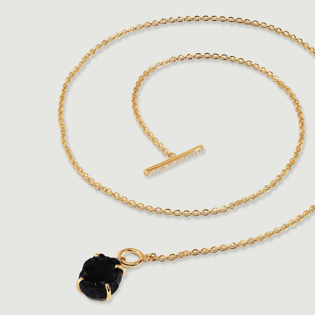 A yellow gold vermeil necklace is laid out in sweeping loops with its black obsidian pendant near the lower edge of a gray surface.