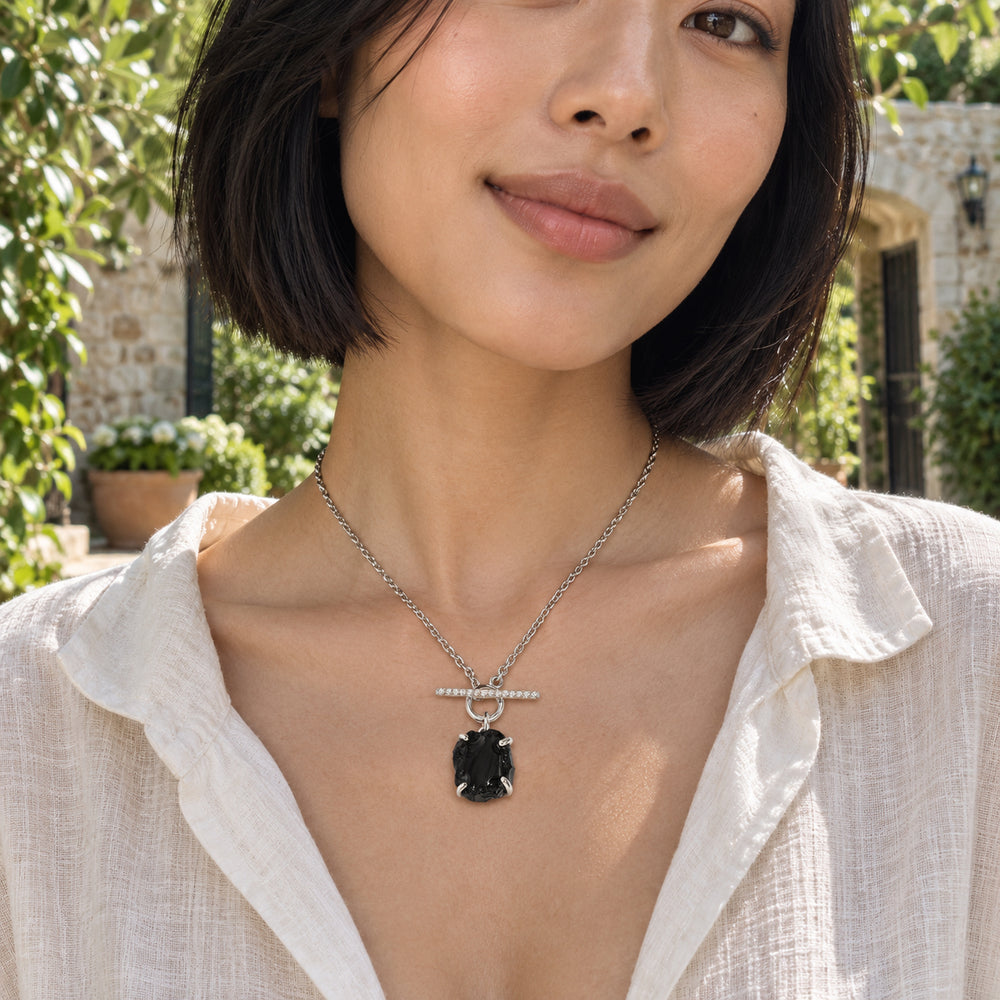 A woman with a black bob wears a silver necklace with a black obsidian pendant and sparkling front bar in a leafy stone courtyard.