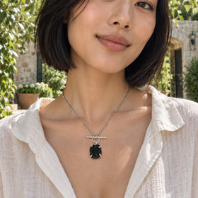A woman with a black bob wears a silver necklace with a black obsidian pendant and sparkling front bar in a leafy stone courtyard.