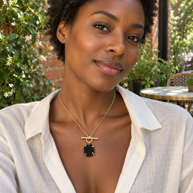 A woman in a white linen shirt wears a yellow gold vermeil necklace with a raw black obsidian pendant at an outdoor cafe.