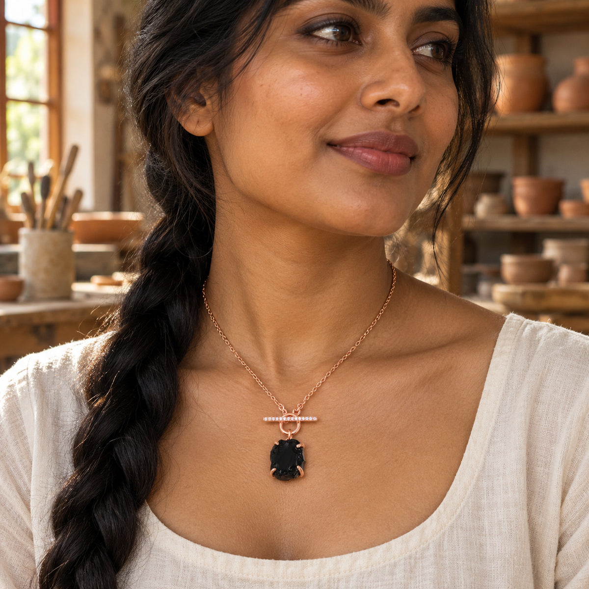 A woman with a long dark braid wears a rose gold vermeil chain and black obsidian pendant in a sunlit ceramics workshop.