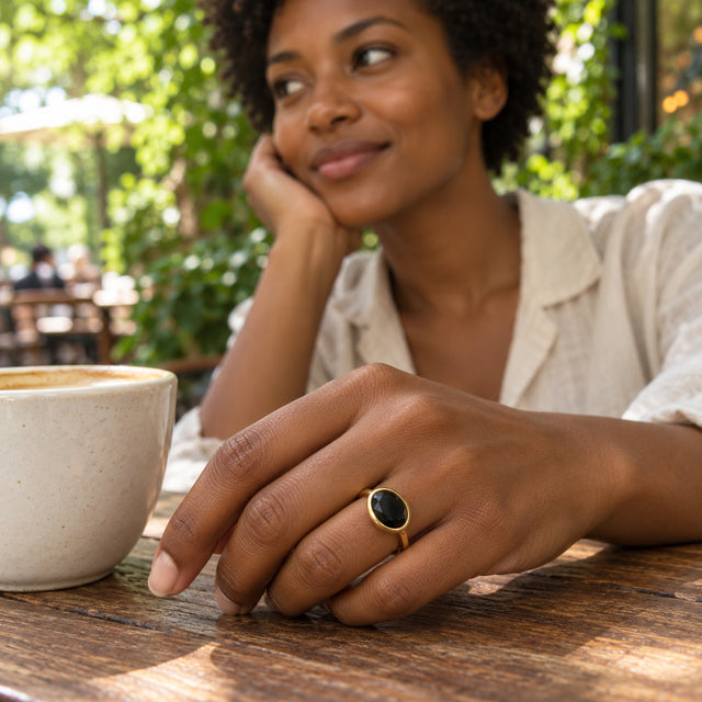 A woman with short curls wears the yellow gold vermeil black stone ring beside a ceramic cup at an outdoor cafe.