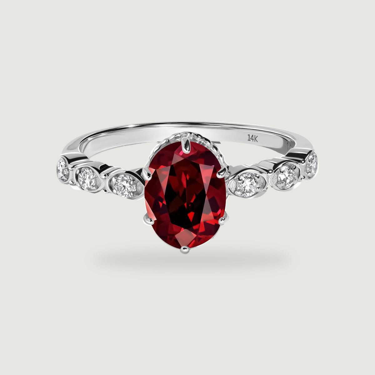 A white gold ring with an oval red garnet and diamond shoulders rests above a soft shadow.