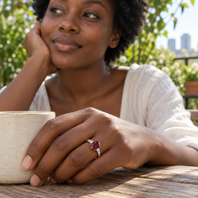 A rose gold garnet ring is worn on a hand resting beside a ceramic cup on a leafy terrace.