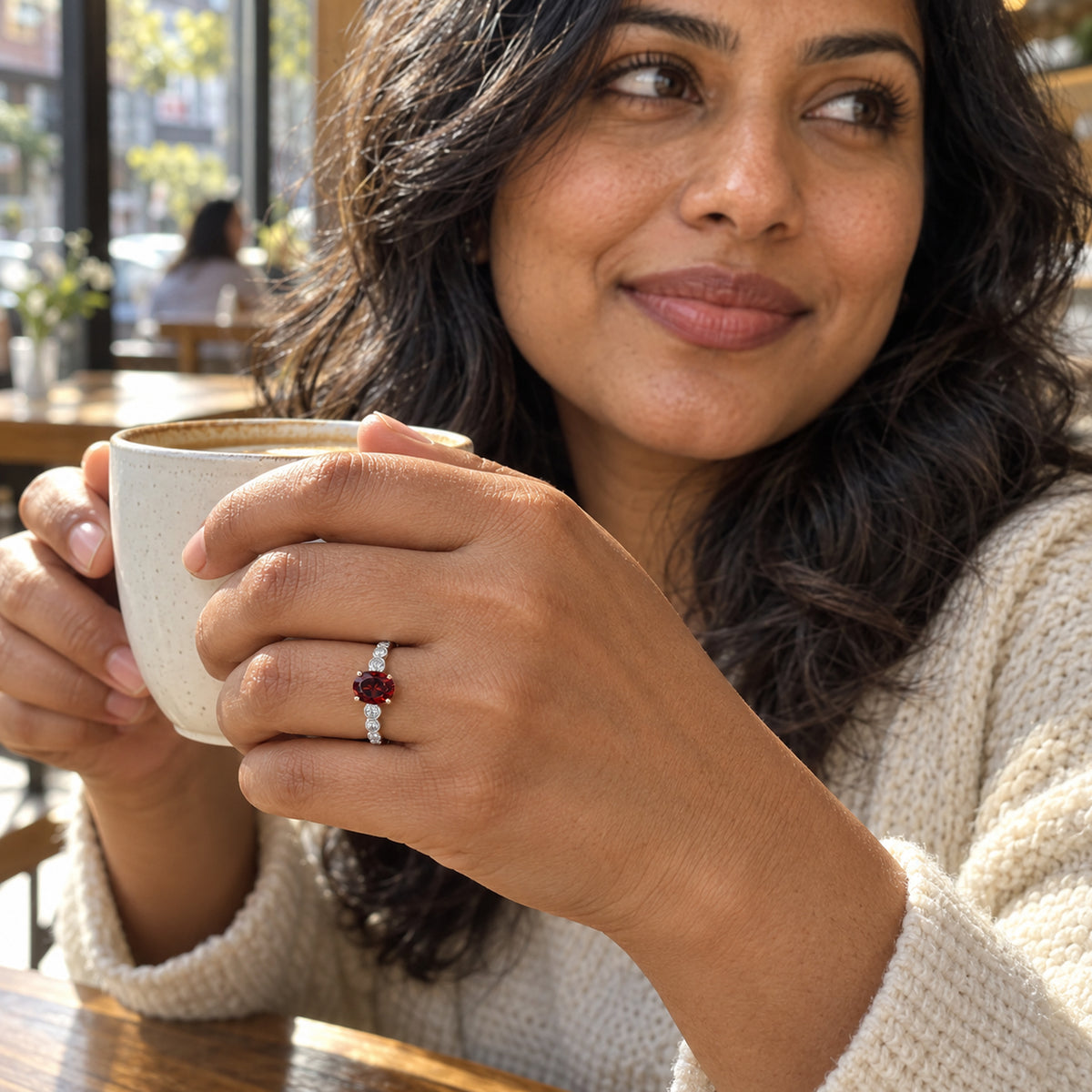 A white gold garnet and diamond ring is worn on a hand holding a ceramic cup in a cafe.