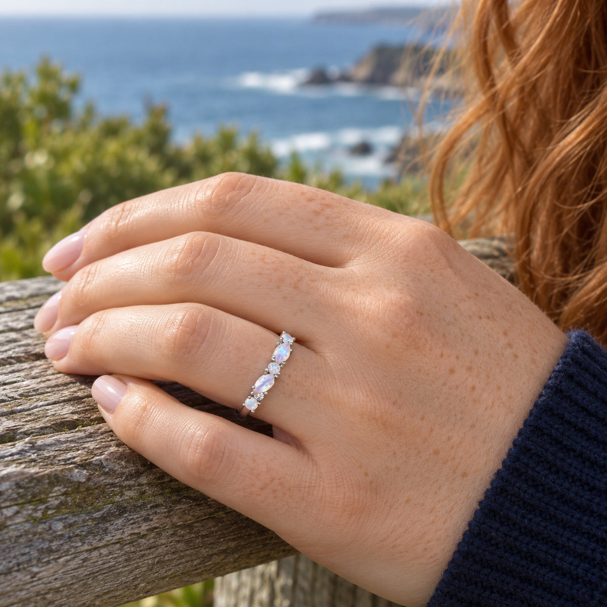 A silver gemstone ring is worn on a freckled hand resting on a coastal wooden rail.