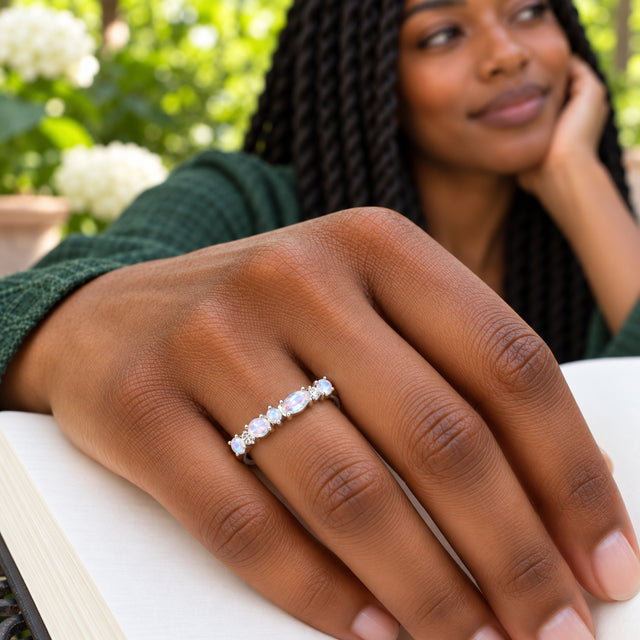 A silver gemstone ring is worn on a brown hand resting on an open book in a leafy garden.