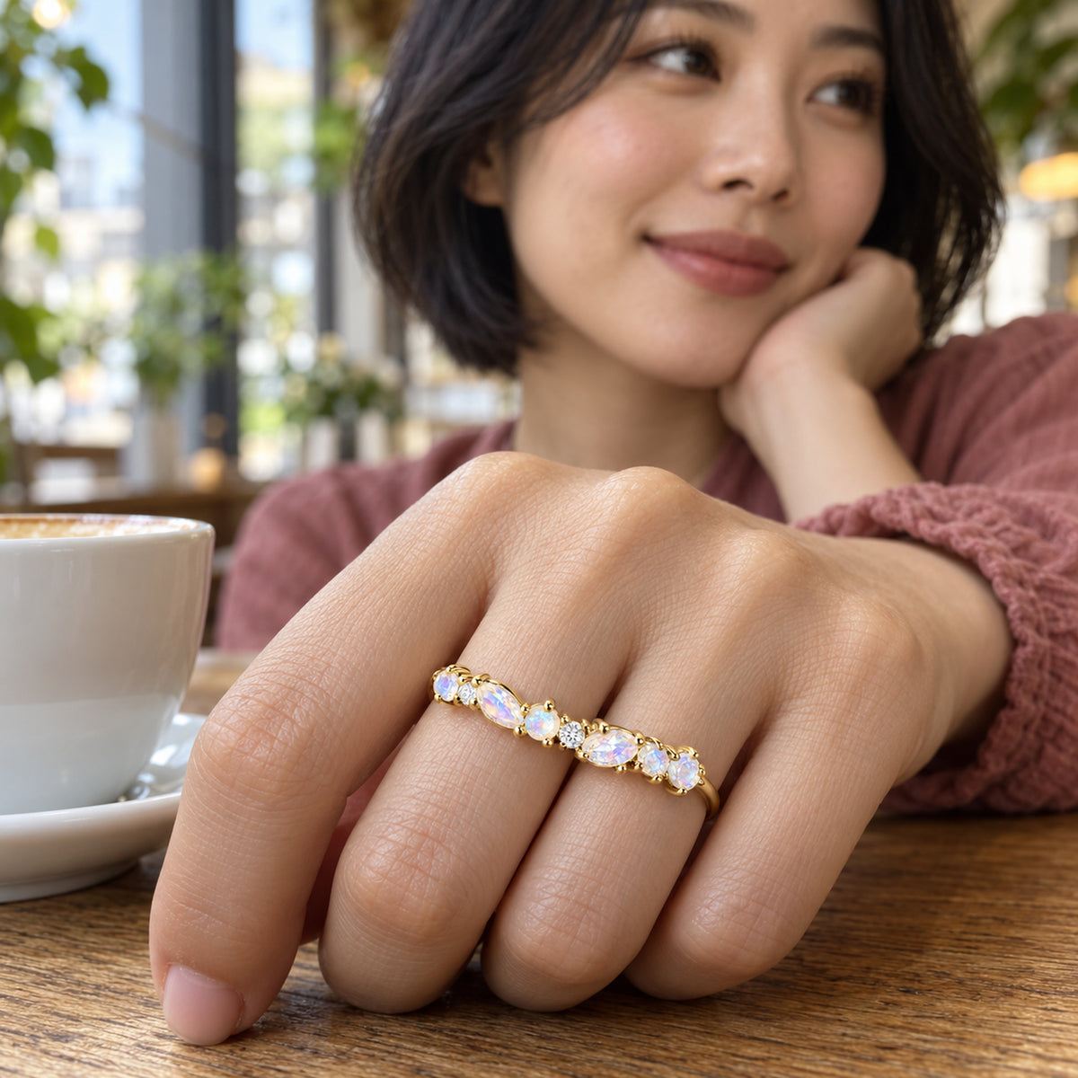 A yellow gold ring with pale stones is shown on a hand beside a white cup on a cafe table.