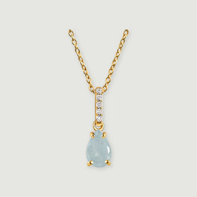 A pale blue pear-shaped pendant hangs below a diamond-set bail on a yellow gold necklace against light gray.