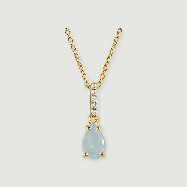 A pale blue pear-shaped pendant hangs below a diamond-set bail on a yellow gold necklace against light gray.