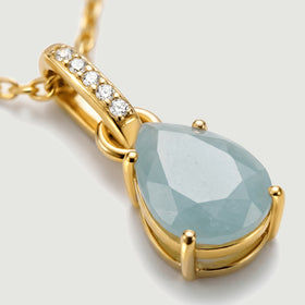 Four yellow gold prongs hold a faceted blue stone beside the diamond-set bail in this close-up.