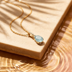 A yellow gold necklace lies across a pale stone slab with its blue pendant and curved chain in sunlight.