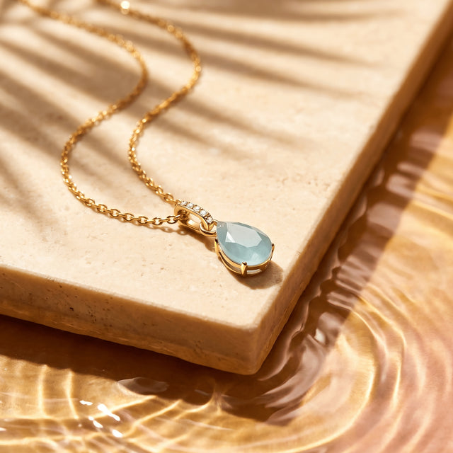 A yellow gold necklace lies across a pale stone slab with its blue pendant and curved chain in sunlight.
