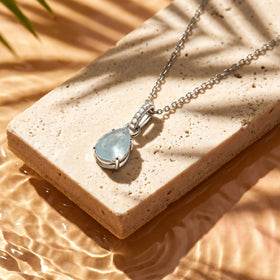 A silver necklace rests diagonally on a cream stone slab with the blue pear-shaped pendant near the lower center.