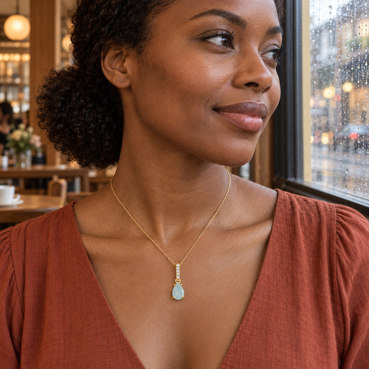A yellow gold necklace with a blue drop pendant is worn above a rust-colored neckline beside a rain-covered cafe window.
