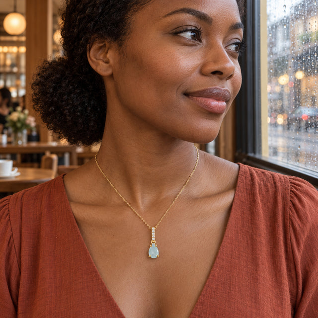 A yellow gold necklace with a blue drop pendant is worn above a rust-colored neckline beside a rain-covered cafe window.