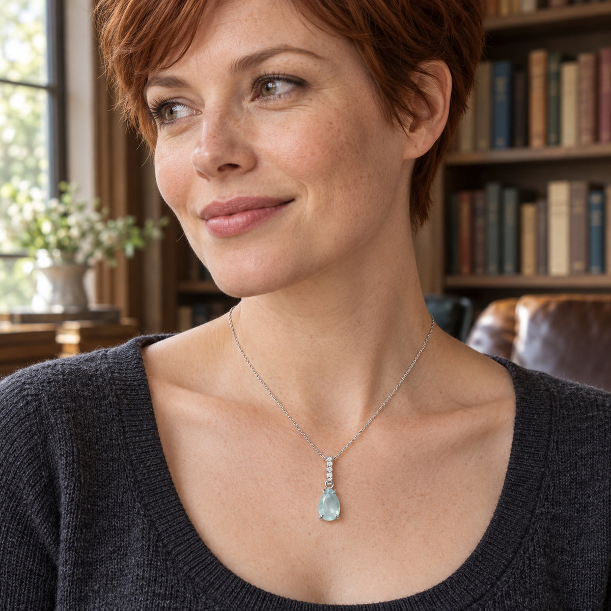 A blue pear-shaped pendant on a silver chain rests over a charcoal knit neckline in a room with bookshelves.