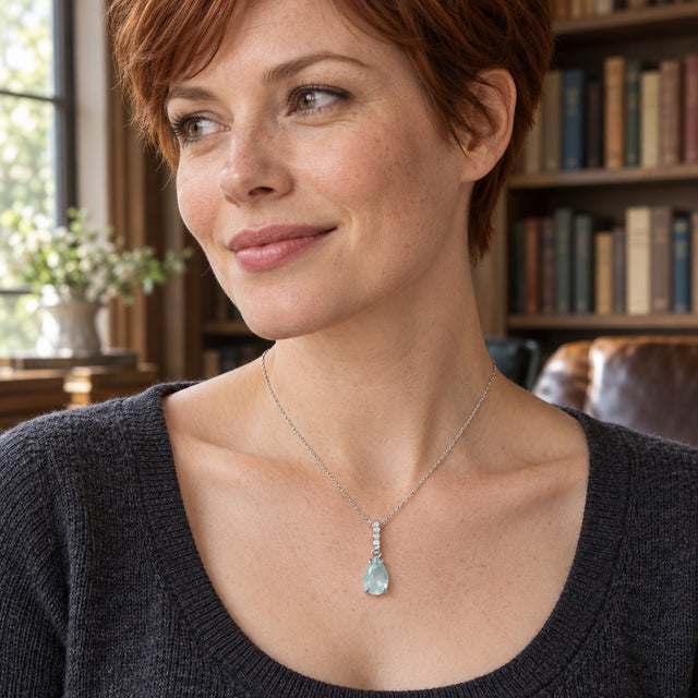 A blue pear-shaped pendant on a silver chain rests over a charcoal knit neckline in a room with bookshelves.