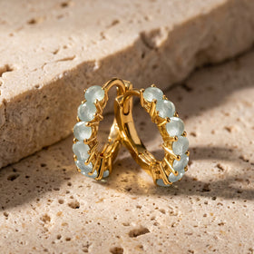 A pair of yellow gold-toned aquamarine hoops stands together on a beige stone tabletop.