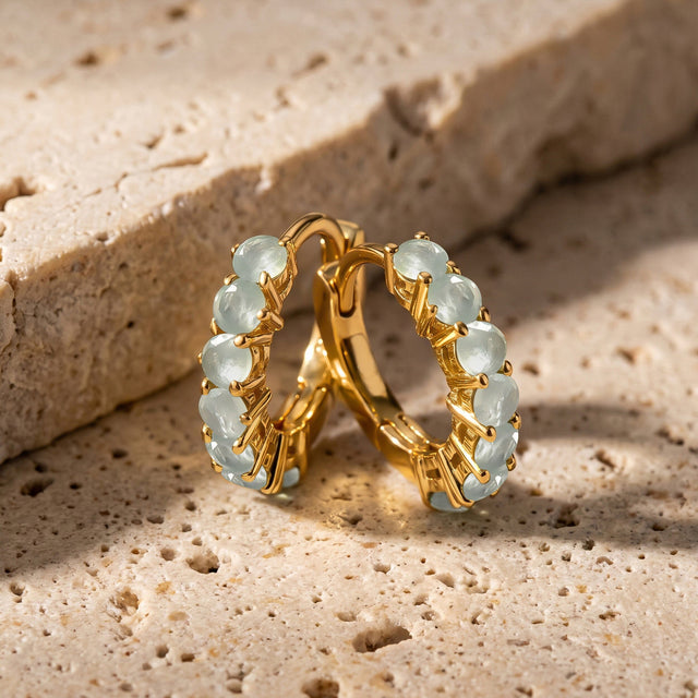 A pair of yellow gold-toned aquamarine hoops stands together on a beige stone tabletop.