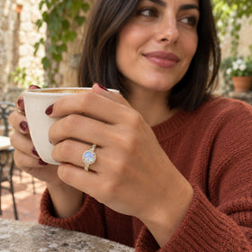A woman with burgundy nails wears the yellow gold moonstone ring while holding a cream ceramic cup in a courtyard.