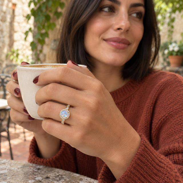 A woman with burgundy nails wears the yellow gold moonstone ring while holding a cream ceramic cup in a courtyard.