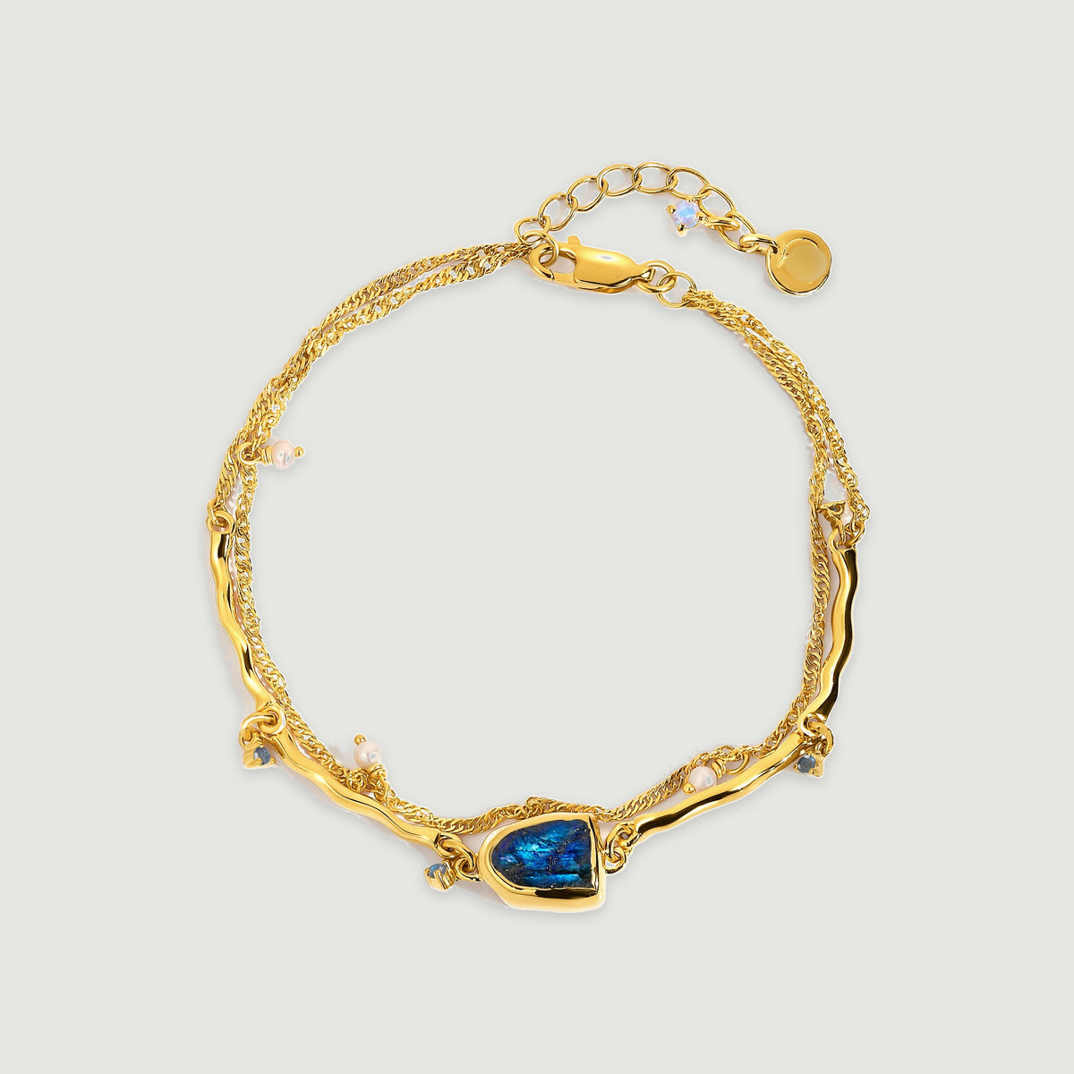 A yellow gold bracelet with a blue labradorite centerpiece, white pearl charms, double chains, and an extender rests on a light gray surface.