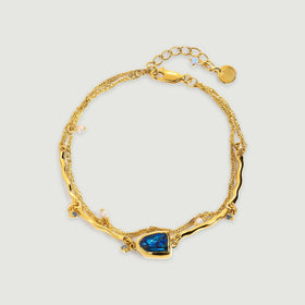 A yellow gold bracelet with a blue labradorite centerpiece, white pearl charms, double chains, and an extender rests on a light gray surface.