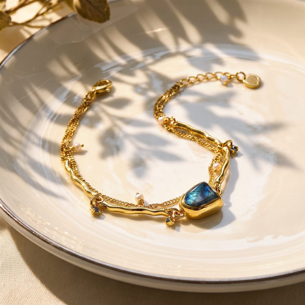 The yellow gold bracelet lies open on a cream ceramic plate, showing its blue stone, pearl charms, and lobster clasp.