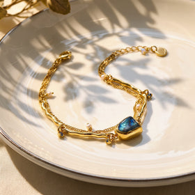 The yellow gold bracelet lies open on a cream ceramic plate, showing its blue stone, pearl charms, and lobster clasp.