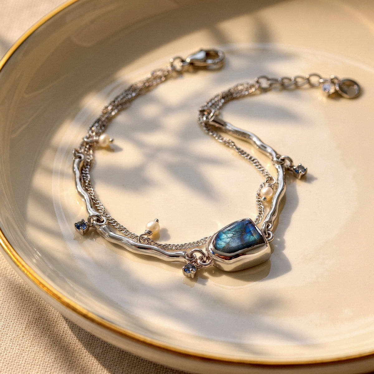 An open silver bracelet with a blue labradorite centerpiece and white pearl charms rests on a ceramic plate in soft window light.