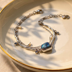 An open silver bracelet with a blue labradorite centerpiece and white pearl charms rests on a ceramic plate in soft window light.