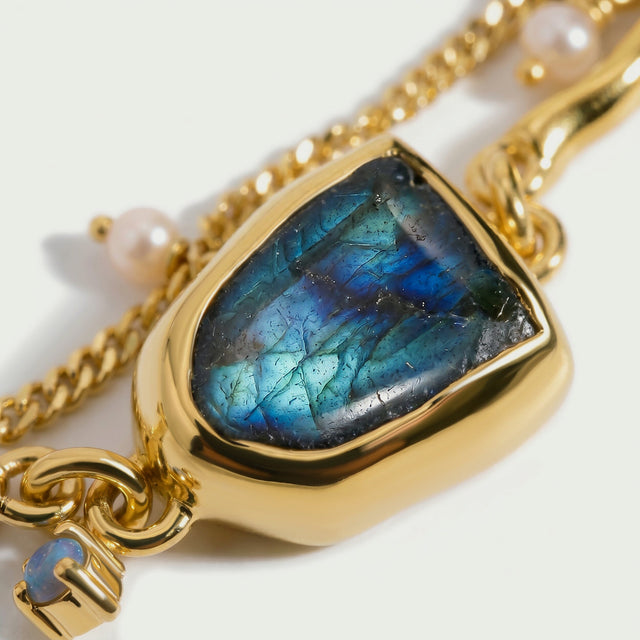 A close view shows the blue and green texture of labradorite inside a polished yellow gold bezel beside two pearl charms.