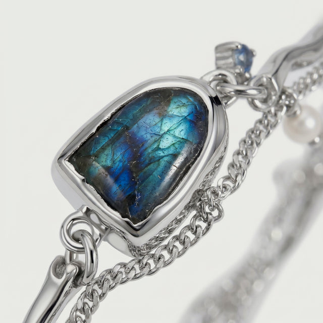 Blue and turquoise veins cross the labradorite surface in a silver bezel, with double chain links and a pearl visible nearby.