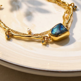 The raised yellow gold bezel and blue labradorite are seen at an angle beside a curved polished bar, a pearl charm, and fine chain links.
