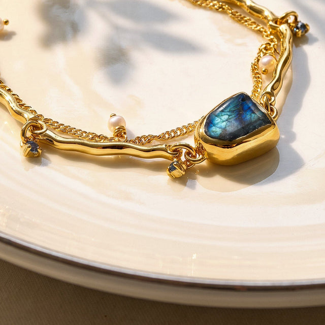 The raised yellow gold bezel and blue labradorite are seen at an angle beside a curved polished bar, a pearl charm, and fine chain links.