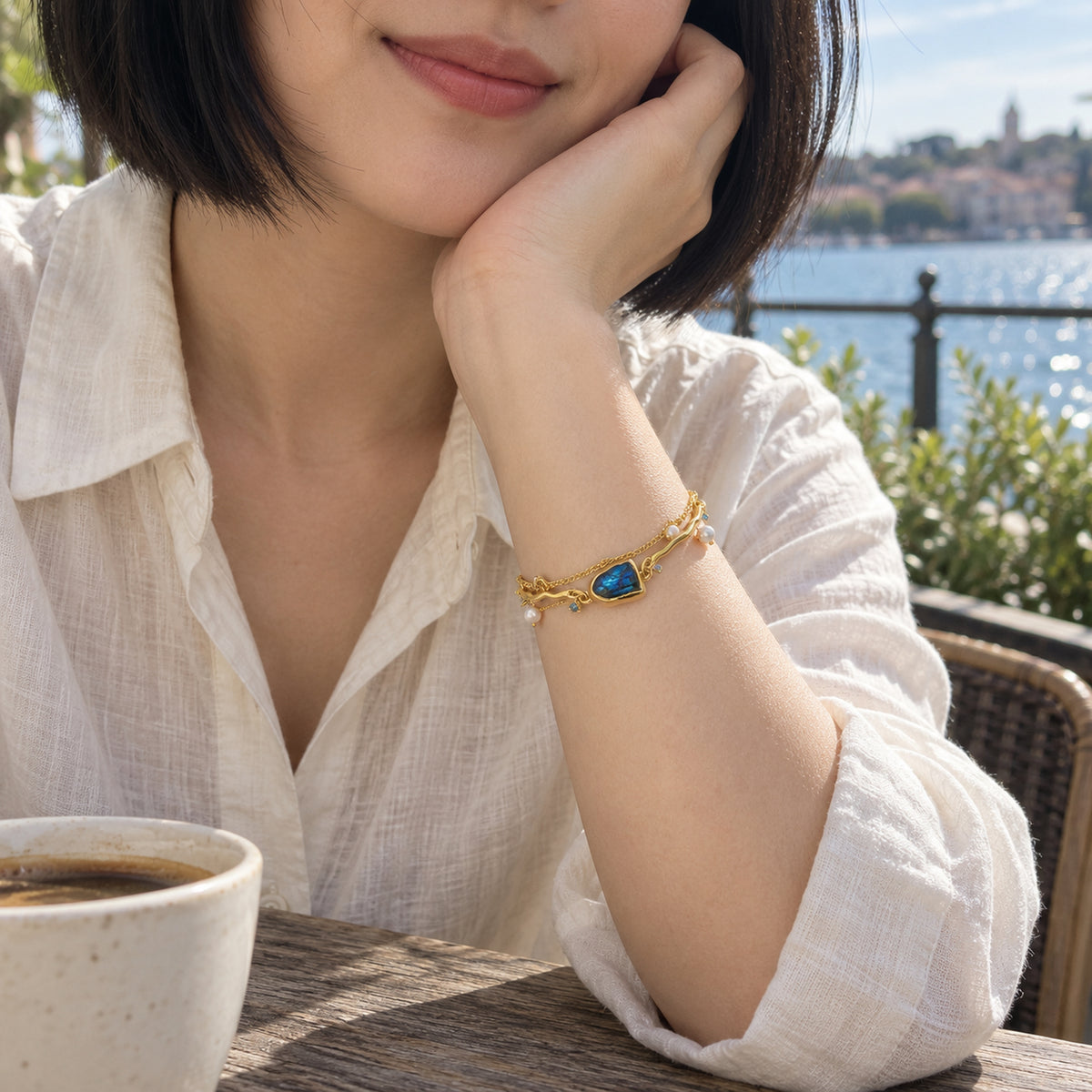 A yellow gold bracelet with a blue labradorite center and white pearl charms circles a wrist beside an ivory linen shirt at a waterfront cafe.