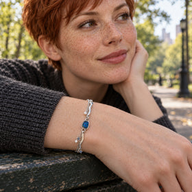A silver bracelet with a blue stone, curved polished bars, and white pearl charms is worn above a dark knitted sleeve beside a green park bench.