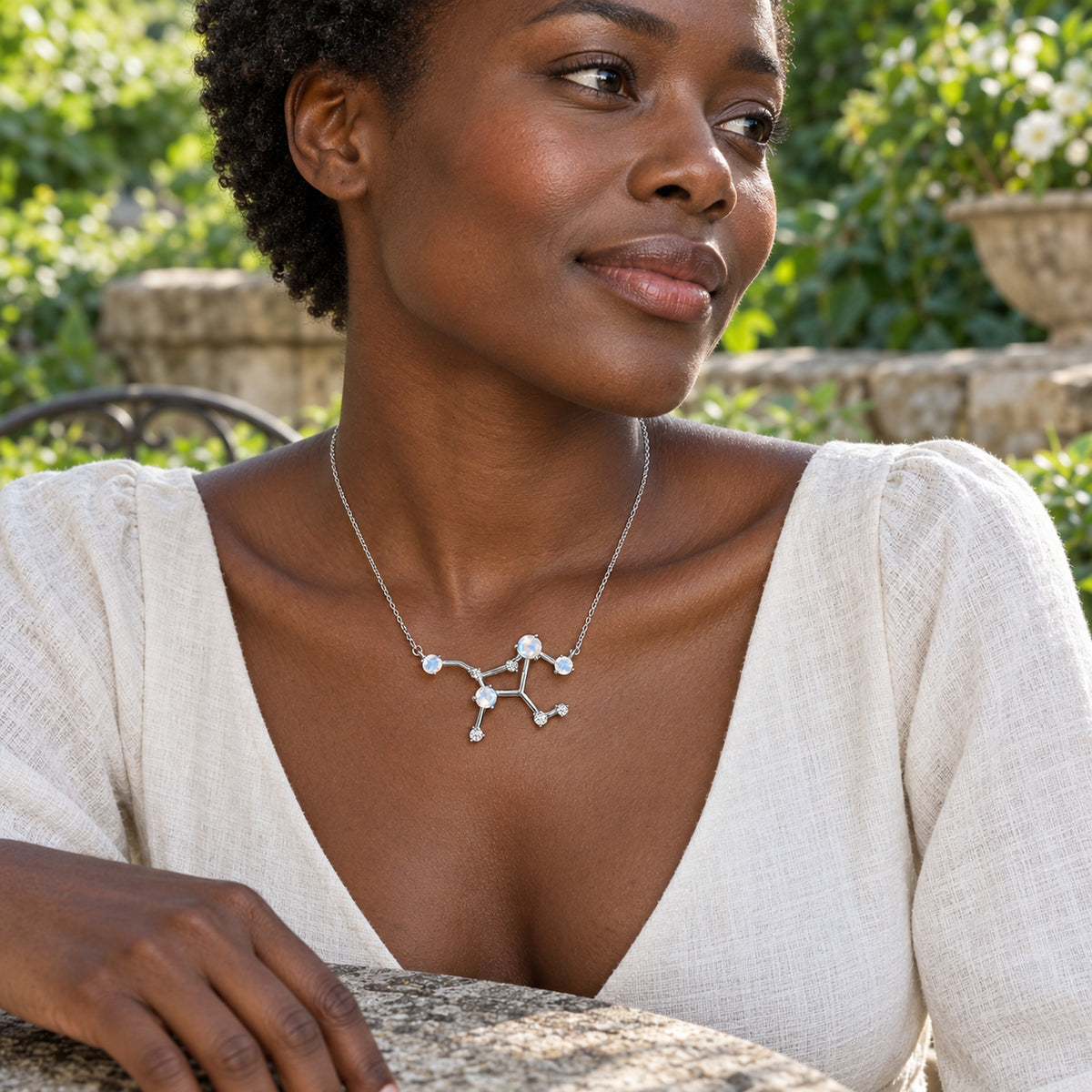 A woman in a cream V-neck blouse wears the silver moonstone necklace at a garden table.