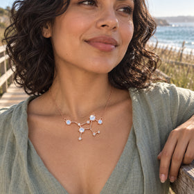 A woman with curly dark hair wears the rose gold pendant and a sage linen top beside a seaside boardwalk.