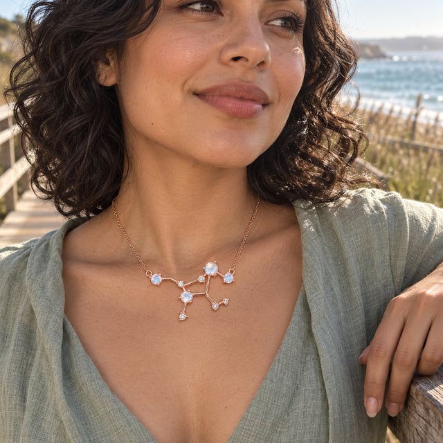 A woman with curly dark hair wears the rose gold pendant and a sage linen top beside a seaside boardwalk.