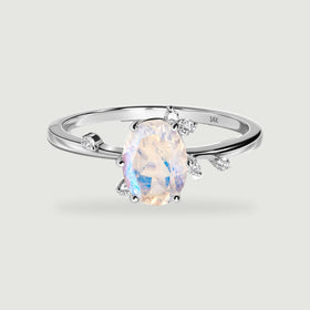 A white gold ring shows a pale oval moonstone with blue reflections and round diamond accents around the setting.