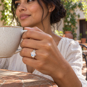 A white gold moonstone ring appears on a hand holding a ceramic tea cup in a leafy courtyard.