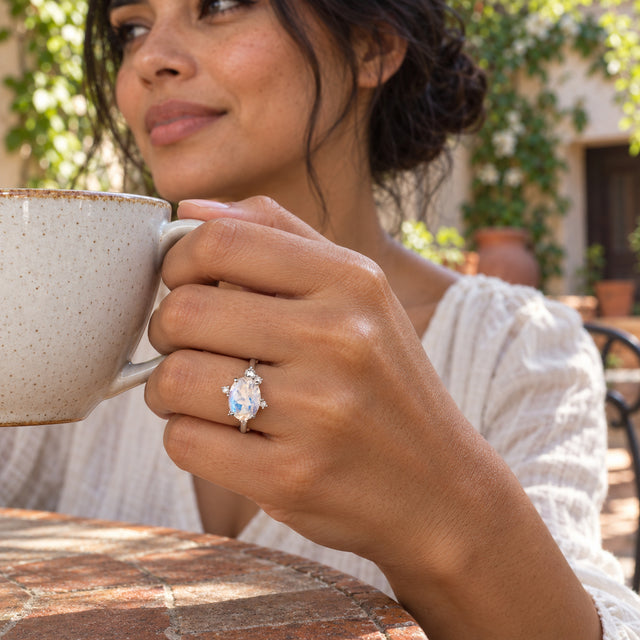 A white gold moonstone ring appears on a hand holding a ceramic tea cup in a leafy courtyard.