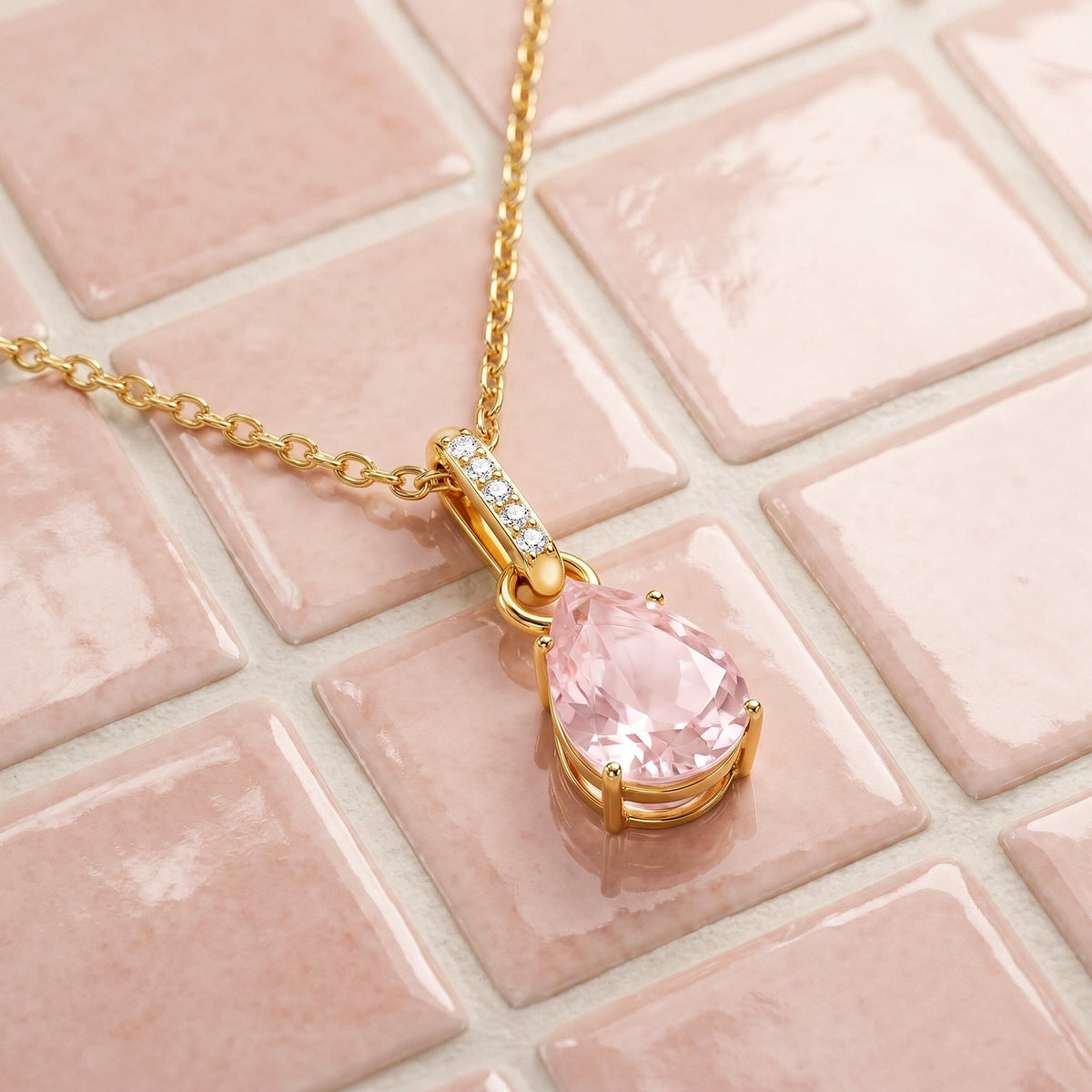 A yellow gold necklace with a faceted pink pear-shaped pendant lies diagonally across pale pink ceramic tiles.