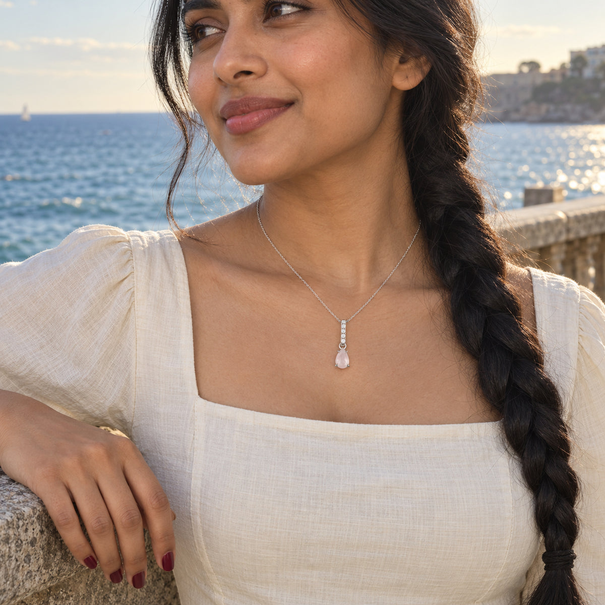 A silver rose quartz necklace rests above a cream square neckline as a woman with a long braid stands beside the sea.