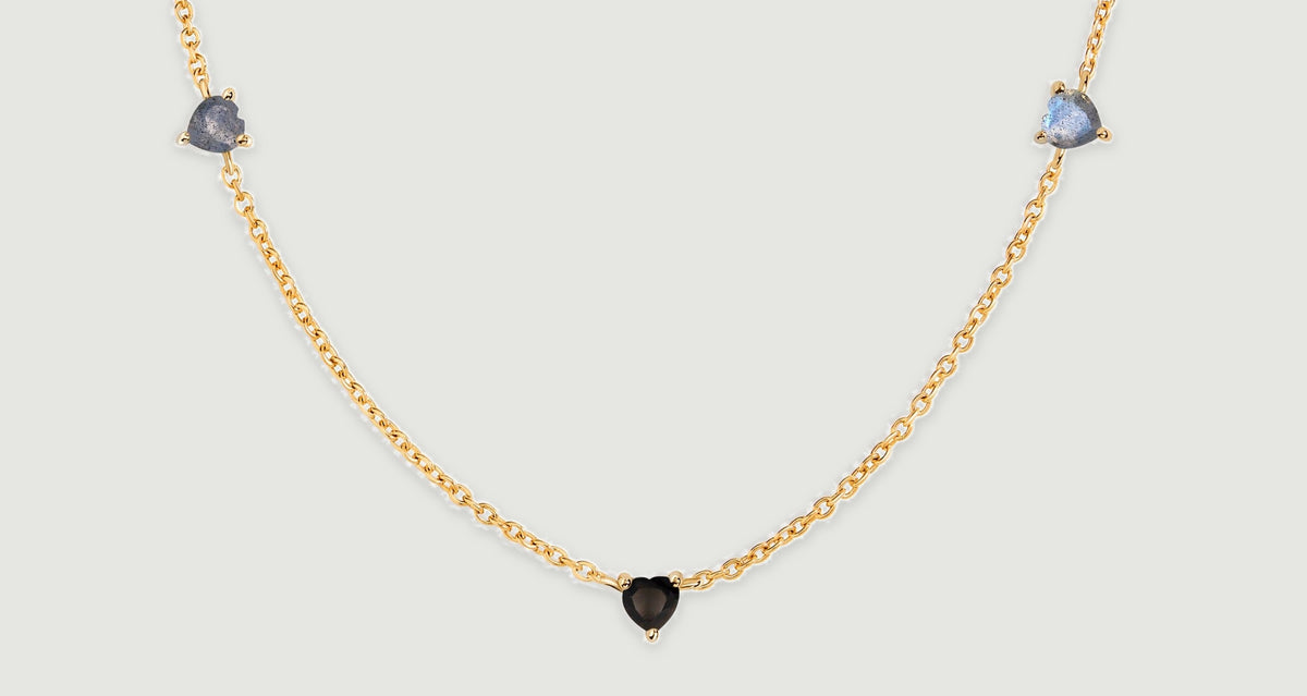 Two blue-gray heart-shaped stones flank a lower black heart along a yellow gold chain laid on a gray surface.