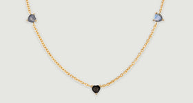 Two blue-gray heart-shaped stones flank a lower black heart along a yellow gold chain laid on a gray surface.