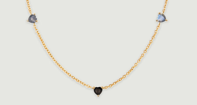 Two blue-gray heart-shaped stones flank a lower black heart along a yellow gold chain laid on a gray surface.